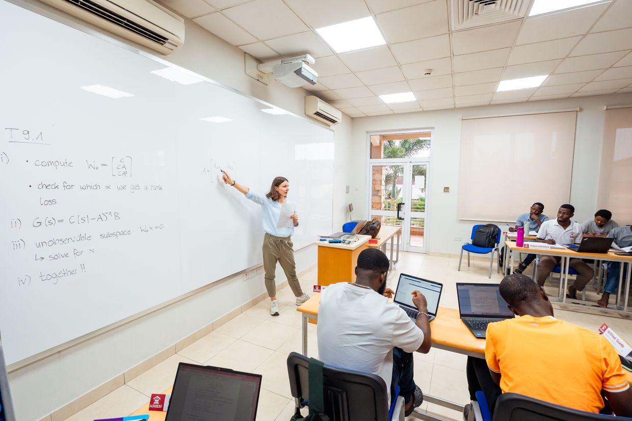 Professor stands at the blackboard and explains something, the students sit in a semi-circle and listen.